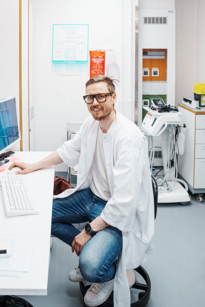 Käsikirurgian lääkäri Anttila istuu tietokonepäätteen äärellä leikkaussalissa ja katsoo kameraan. Paikallispuudutuksessa tehtävä käsikirurgia säästää resursseja.